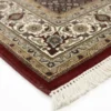 Detailed corner view of a burgundy Indian wool rug showing the ornate cream border and clean white hand-bound fringe.