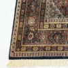 Detailed corner view of a dark Indian wool rug showing the wide ornate floral border and clean white hand-bound fringe.