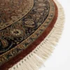 Detailed edge view of a circular burgundy Indian rug showing the ornate navy border and authentic white hand-bound fringe.