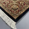 Detailed corner view of a black Indian rug showing the ornate gold-tan botanical border and clean white hand-bound fringe.