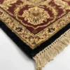 Detailed corner view of a black Indian rug showing the ornate burgundy botanical border and clean white hand-bound fringe.