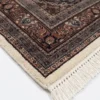 Detailed corner view of an Indian wool rug showing the ornate plum and tobacco brown botanical border and clean white hand-bound fringe.