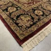 Detailed corner view of a red Indian rug showing the ornate black botanical border with gold accents and clean white hand-bound fringe.