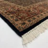 Detailed corner view of a tobacco brown Indian rug showing the ornate rust botanical border and clean white hand-bound fringe.