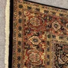 Close-up corner view of a square tobacco and rust Indian wool rug showing the hand-bound dark edges and thick cream fringe.
