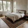 Airy minimalist bedroom with a walnut bed frame and neutral linens, featuring a square tobacco Indian rug at the foot of the bed.