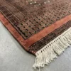 Detailed corner view of a coral Pakistan Bokhara rug showing the multi-layered geometric border and clean white hand-bound fringe.