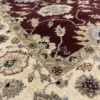 Detailed corner view of a burgundy Chobi rug showing the wide ornate ivory botanical border and clean white hand-bound fringe.