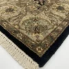 Detailed corner view of a black Indian rug showing the ornate olive-gold botanical border and clean white hand-bound fringe.