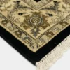 Detailed corner view of a black Indian rug showing the ornate ivory and gold botanical border and clean white hand-bound fringe.