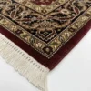 Detailed corner view of a red Indian rug showing the ornate gold and ivory botanical border and clean white hand-bound fringe.