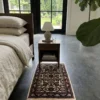 Serene modern bedroom with dark concrete floors and a walnut nightstand, featuring a small ivory and sage hand-knotted Indian wool rug placed at the foot of the nightstand.