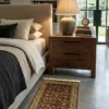 Peaceful modern bedroom featuring a walnut nightstand and light linens, with a small burgundy and gold Indian wool rug providing a warm tactile accent on gray floors.