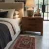 Peaceful modern bedroom featuring a walnut nightstand and light linens, with a small terracotta and navy Gholtogh Persian rug on light gray polished concrete floors.