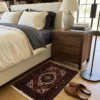 Peaceful modern bedroom featuring a walnut nightstand and light linens, with a small blue and red Gholtogh Persian rug providing a warm tactile accent on polished tan concrete floors.