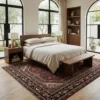 Peaceful primary bedroom with a mid-century walnut bed frame and beige linens, anchored by an intricate red Hosinabad Persian rug under arched windows.
