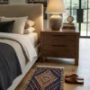 Minimalist bedroom featuring a walnut nightstand and light linens, with a small navy and orange Qashqai wool rug providing a tactile accent on a gray floor.