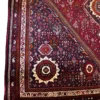 Technical close-up corner view of a plum and red Iranian rug showing the intricate ivory guard border and short, hand-tied fringe.