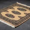 An angled perspective view of a small gold Pakistan Bokhara rug on a dark gray floor, showing the rug's thickness and vibrant color.