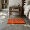 Serene modern bedroom featuring a walnut nightstand and light linens, with a small orange tufted wool rug providing a warm color pop on a polished concrete floor.