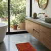 Minimalist bathroom with a floating walnut vanity and concrete counters, featuring a small orange tufted rug as a functional accent on a gray floor.