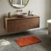 Minimalist bathroom with a floating walnut vanity and concrete counters, featuring a small orange tufted rug as a functional accent on a gray floor.
