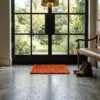 Bright entryway with a leather bench and glass doors, featuring a small sunset-orange tufted rug centered on a gray concrete floor.