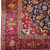 Close-up corner view of a Persian Kashan rug highlighting the dense red floral border, navy spandrels, and flat hand-bound edge finish.