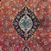 Close-up macro shot of the central diamond medallion on an Iranian Kashan wool rug, showing the high-density red, navy blue, and sky blue curvilinear floral details.