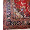 Technical corner view of a red and navy Persian Sabzevar rug highlighting the multi-guard botanical border and flat hand-wrapped edge finish.